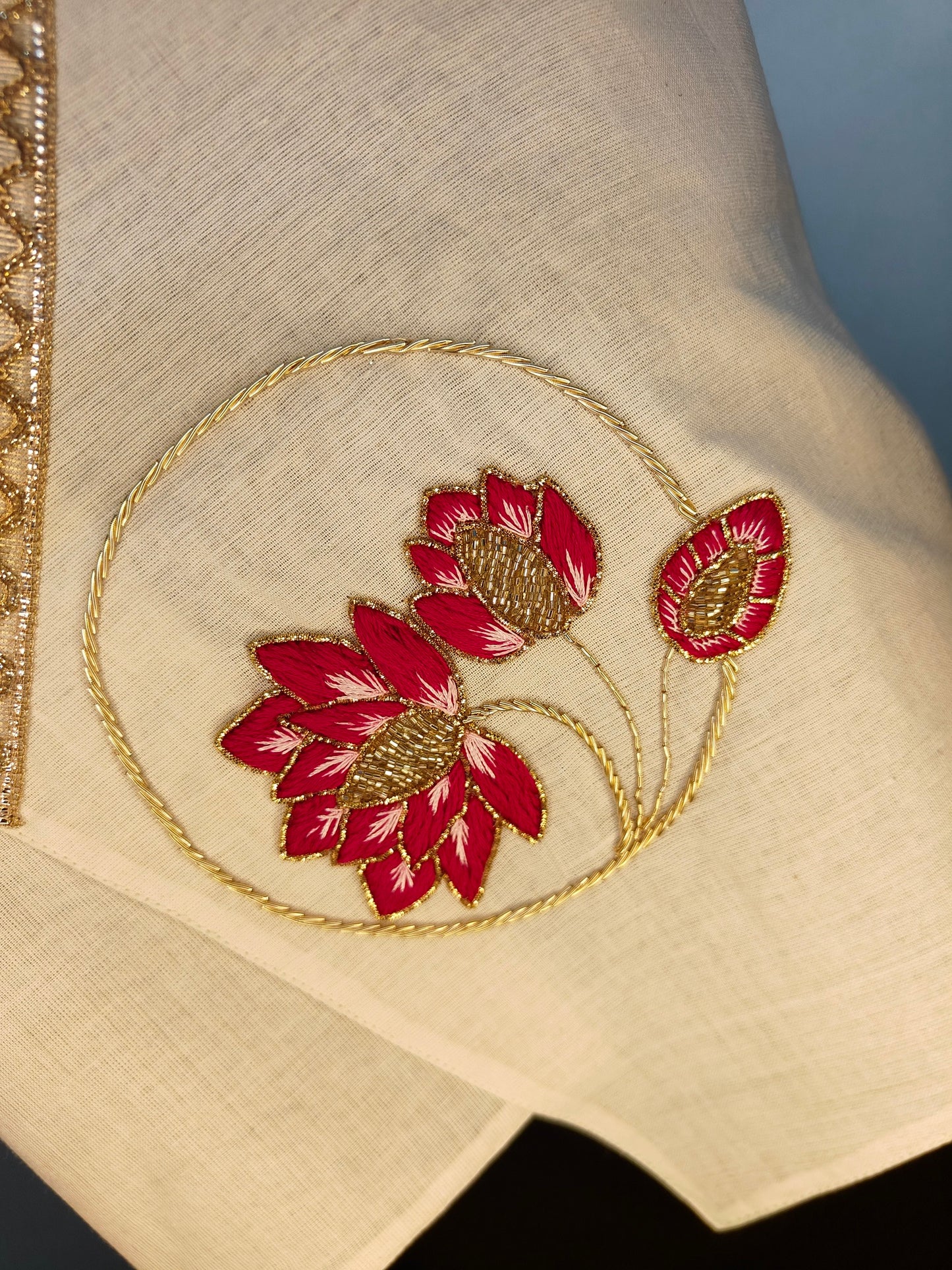 Close-up of embroidered fabric with red flowers and gold detailing on a beige background.